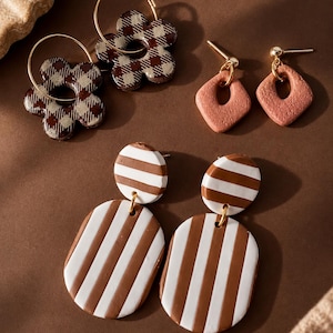 May include: Three pairs of earrings: one pair of gold hoop earrings with brown and beige checkered flower charms, one pair of textured terracotta diamond-shaped stud earrings, and one pair of brown and white striped oval drop earrings.