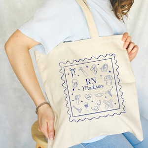 May include: Cream-colored tote bag with a blue illustrated design of medical tools and the text "RN + Madison" inside a square border. The bag has a simple design with long straps.