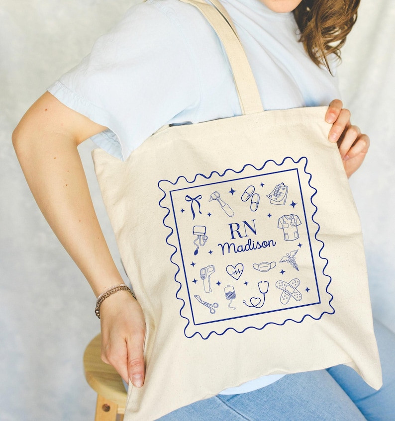 May include: Cream-colored tote bag with a blue illustrated design of medical tools and the text "RN + Madison" inside a square border. The bag has a simple design with long straps.