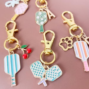 May include: A collection of gold-colored keychains with various charms. Charms include pickleball paddles in blue, pink, and green, a paw print, cherries, and the letter "A". The keychains have gold clasps and key rings.