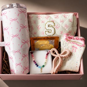 May include: A pink gift box filled with a white tumbler with pink bow accents, a bow-shaped hair clip, a beaded necklace, a cosmetic bag, socks, and eye masks. The tumbler and cosmetic bag have bow patterns.
