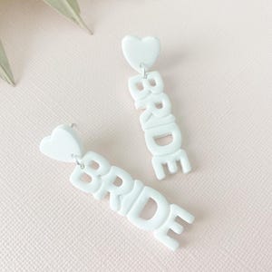 May include: A pair of white BRIDE earrings. Each earring features a heart-shaped stud and the word "BRIDE" in capital letters. The earrings are made of a solid, opaque material and hang vertically.