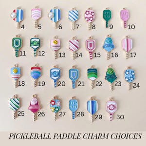 May include: Thirty different pickleball paddle charms with various designs and colors. The charms are made of metal and have a gold finish. Some of the designs include flowers, stripes, polka dots, and the words "Let's Play".