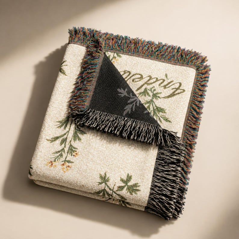 Pode incluir: Uma manta tecida com uma base de cor creme e um padr&atilde;o bot&acirc;nico de folhas verdes e pequenas flores amarelas. A manta tem um canto dobrado que revela um verso preto e &eacute; finalizada com uma borda de franjas multicoloridas.