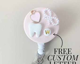 May include: Pink retractable badge holder with a white tooth, a pink heart, a pearl bow, and a gold letter "W".