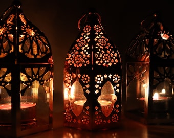 Peut inclure: Trois lanternes en métal ornées de motifs complexes, chacune contenant une bougie allumée à l'intérieur. Les lanternes sont de couleur cuivre chaud et ont un style marocain traditionnel.
