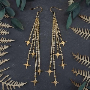 Può includere: Orecchini pendenti color oro con più catene e ciondoli a forma di stella. Gli orecchini hanno una chiusura a gancio e sono mostrati su uno sfondo scuro con fogliame verde e oro. Un accessorio a tema celeste.