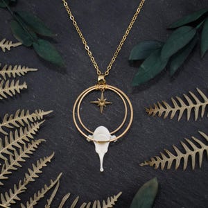 Può includere: Una collana d'oro con un ciondolo bianco a forma di lacrima che cola. Il ciondolo è circondato da due cerchi dorati, e una stella dorata è centrata all'interno dei cerchi.