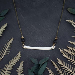 Puede incluir: Un collar único con un colgante de hueso blanco suspendido de una delicada cadena dorada. El collar incluye detalles de cuentas negras y está rodeado de hojas de helecho sobre un fondo oscuro.