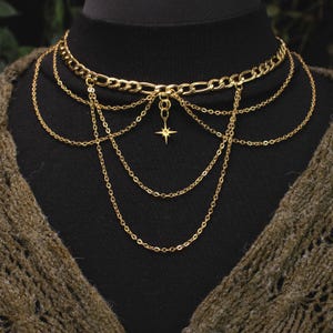 Puede incluir: Collar dorado en capas con una cadena tipo gargantilla y múltiples cadenas delicadas en cascada. Un pequeño colgante de estrella cuelga del centro. El collar se muestra sobre una superficie negra.