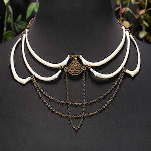 May include: A distinctive necklace with a bone-like design, featuring three curved white elements. It has an antique brass-coloured centrepiece and multiple hanging chains. The necklace is displayed on a black mannequin.