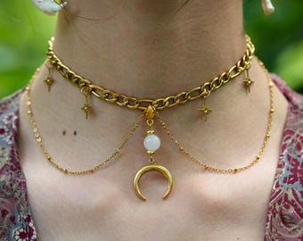 May include: A gold-toned chain choker necklace featuring four small star-shaped charms hanging from the main links. A central drop element consists of a small white spherical bead suspended above a gold-toned crescent moon pendant on a thin chain.
