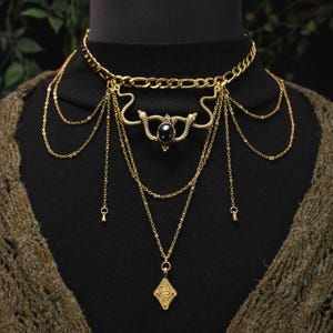 Op de afbeelding: Goudkleurige ketting met een choker-stijl ketting, waterval kettingen en een centrale hanger met een zwarte steen en slangenmotief. Een ruitvormige bedel hangt aan de langste ketting. De ketting wordt op een zwarte achtergrond getoond.