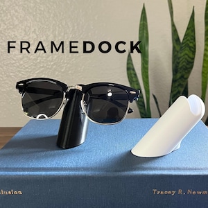 May include: Black sunglasses with silver detailing are displayed on a black stand. A white stand is next to it. The word FRAMEDOCK is printed in large black letters above the sunglasses. The items are on a blue book.