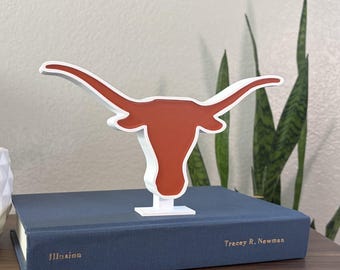 May include: A white and burnt orange Longhorn steer head sculpture. The sculpture is displayed on a blue book with the title "Illusion" and the author's name, Tracey R. Newman. The sculpture is on a wooden surface.