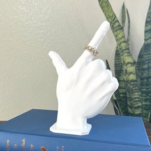 May include: White ceramic hand ring holder with a gold ring with clear stones on the index finger. The hand holder is sitting on a blue book with the title "Where the Crawdads Sing" by Delia Owens.