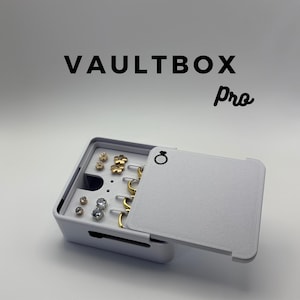 May include: A white square jewelry storage box labeled VAULTBOX Pro with a sliding lid featuring a ring icon. The interior contains gold and silver-toned stud earrings and several gold hoop earrings arranged in organized slots and holes.