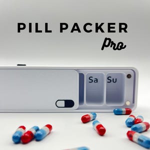 Puede incluir: Un organizador de píldoras blanco con una pantalla digital y el texto "PILL PACKER pro". El organizador tiene siete compartimentos etiquetados con los días de la semana, incluyendo "Sa" para sábado y "Su" para domingo. Hay varias píldoras rojas y azules esparcidas alrededor del organizador.