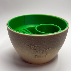 May include: A two-toned bowl with a green interior and a beige exterior. The bowl has a built-in nut cracker and a small cup. The text "The NUT Bowl" is embossed on the side.