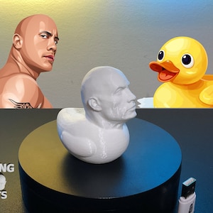 Può includere: Un'anatra di gomma bianca stampata in 3D con un viso umano. L'anatra è seduta su una piattaforma circolare nera. Il testo "ROCKING 3D PRINTS" è visibile sulla piattaforma.