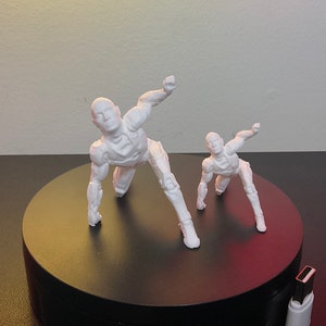 Puede incluir: Dos figuras de acci&oacute;n blancas articuladas en posici&oacute;n agachada, una m&aacute;s grande que la otra. Las figuras est&aacute;n sobre una plataforma negra y circular. Las figuras tienen un dise&ntilde;o futurista y rob&oacute;tico.