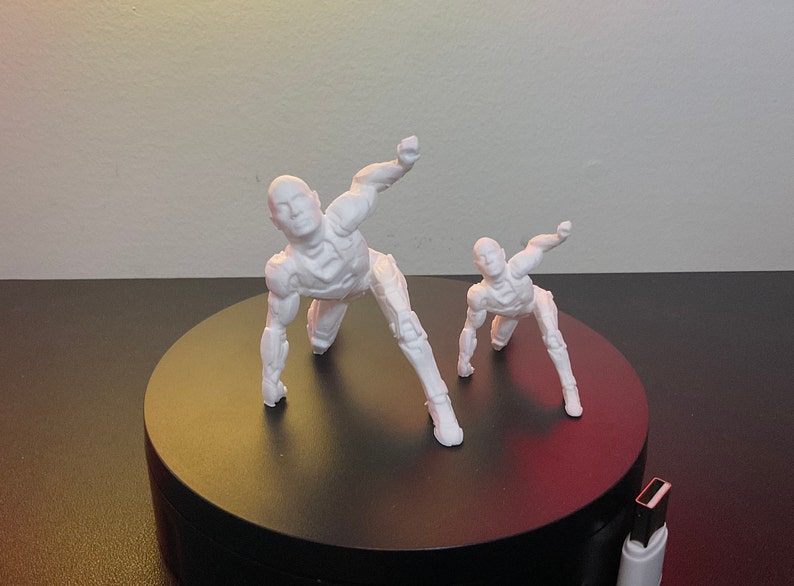 Puede incluir: Dos figuras de acci&oacute;n blancas articuladas en posici&oacute;n agachada, una m&aacute;s grande que la otra. Las figuras est&aacute;n sobre una plataforma negra y circular. Las figuras tienen un dise&ntilde;o futurista y rob&oacute;tico.