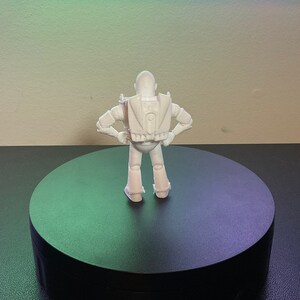 Op de afbeelding: Witte, 3D-geprinte plasticine figuur van een robot met uitgestrekte armen, staand op een ronde zwarte basis.