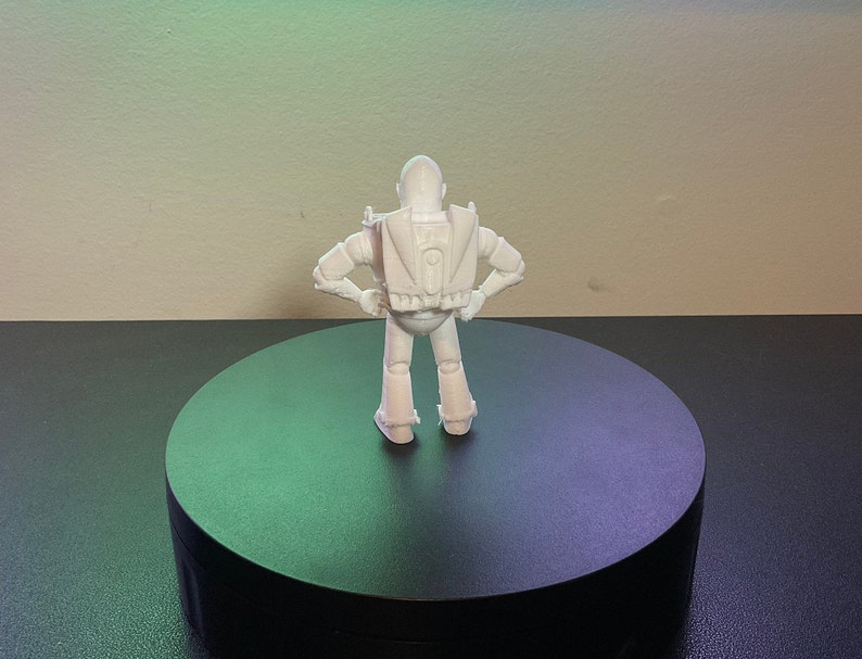 Op de afbeelding: Witte, 3D-geprinte plasticine figuur van een robot met uitgestrekte armen, staand op een ronde zwarte basis.