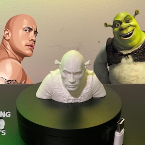 May include: A 3D-printed bust of a bald man with a white vest, placed on a black circular base. The image also features illustrations of Dwayne Johnson and Shrek. The logo "ROCKING 3D PRINTS" is visible in the lower left corner.