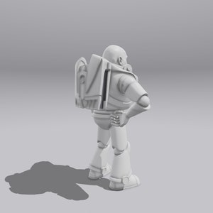 Op de afbeelding: Een wit plastic speelgoedrobot met een rugzak en een helm. De robot staat met zijn rechterarm gebogen in de elleboog en zijn linkerarm recht naar beneden. De robot kijkt naar rechts.