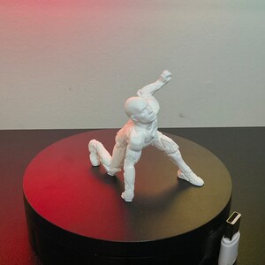 Puede incluir: Una figura blanca de una figura humana en una pose din&aacute;mica, arrodillada con un brazo levantado. La figura est&aacute; sobre una plataforma giratoria negra, con un cable USB visible en primer plano. El fondo es un degradado de rojo y blanco.