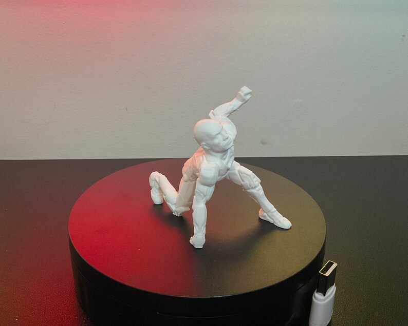 Puede incluir: Una figura blanca de una figura humana en una pose din&aacute;mica, arrodillada con un brazo levantado. La figura est&aacute; sobre una plataforma giratoria negra, con un cable USB visible en primer plano. El fondo es un degradado de rojo y blanco.