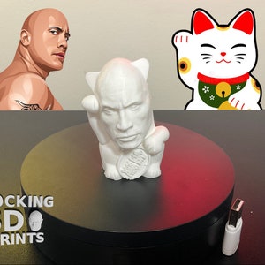 Può includere: Figurina stampata in 3D di colore bianco di un uomo con una somiglianza a Dwayne "The Rock" Johnson, in una posa tradizionale giapponese di Maneki-Neko (gatto che fa cenno). La figurina è seduta su una piattaforma circolare nera con le parole "ROCKING 3D PRINTS" stampate sul davanti.