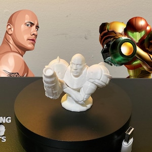 May include: A white 3D-printed bust of Dwayne Johnson in a futuristic suit, displayed on a black rotating platform. The image also features a graphic of Dwayne Johnson and a character from the video game Metroid.
