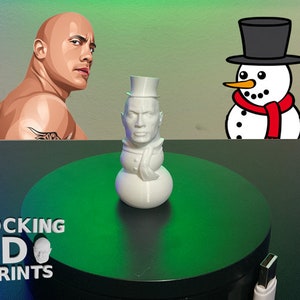 Może przedstawiać: Biała figurka bałwana wydrukowana w 3D z cylindrem i szalikiem. Bałwan ma twarz Dwayne'a "The Rock" Johnsona. Figurka stoi na zielonej, okrągłej platformie z napisem "ROCKING 3D PRINTS" na platformie.