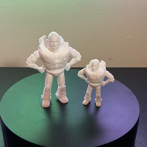 Op de afbeelding: Twee witte 3D-geprinte figuren van Buzz Lightyear uit de Toy Story-franchise. De grotere figuur staat met zijn armen op zijn heupen, terwijl de kleinere figuur met gekruiste armen staat. Beide figuren kijken naar voren.