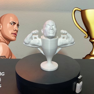 Może przedstawiać: Biały, wydrukowany w 3D trofeum z rzeźbioną głową Dwayne'a "The Rock" Johnsona. Trofeum ma umięśnione ramiona uniesione w pozie flex. Trofeum stoi na czarnym talerzu obrotowym z napisem "ROCKING 3D PRINTS" z boku.