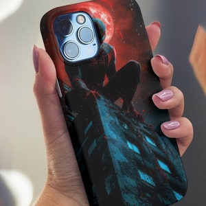 Puede incluir: Una funda de teléfono negra con una luna roja y una figura de superhéroe negra de pie en un edificio. El superhéroe lleva un traje negro con un diseño de araña roja en el pecho.