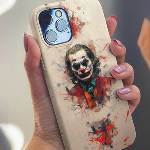 Puede incluir: Una funda de teléfono beige con una ilustración de estilo acuarela del Joker de la película "Joker". El Joker lleva un traje rojo y tiene la cara blanca con lápiz labial rojo y pelo verde. La funda tiene un recorte para la cámara del teléfono.