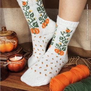 May include: White knit socks with a pumpkin and vine pattern. The socks feature orange pumpkins and green vines, with small orange dots scattered across the white fabric. A cozy autumn theme.