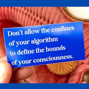 May include: A blue card with white text that reads: "Don't allow the confines of your algorithm to define the bounds of your consciousness." The card is held in front of a pink, textured background.