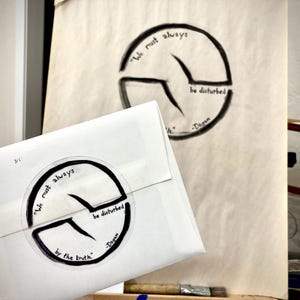May include: A white envelope with a circular design featuring the quote "We must always be disturbed by the truth" and the name "Dogen." A similar design is sketched on a canvas in the background.