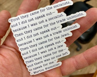 May include: A white sticker with black text quoting Martin Niemöller: "First they came for the socialists... Then they came for me - and there was no one left to speak for me." The sticker is held in a hand.