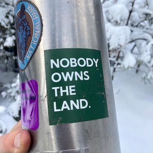 May include: A silver Yeti water bottle with a green sticker that says "Nobody Owns The Land." The bottle has a sticker of a wolf with the words "National Park" on it.