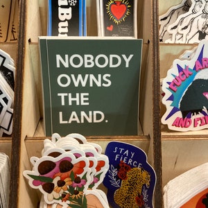 May include: A collection of stickers with various designs, including a green sticker with the text "Nobody owns the land." and a blue sticker with the text "Stay fierce" and a leopard illustration.