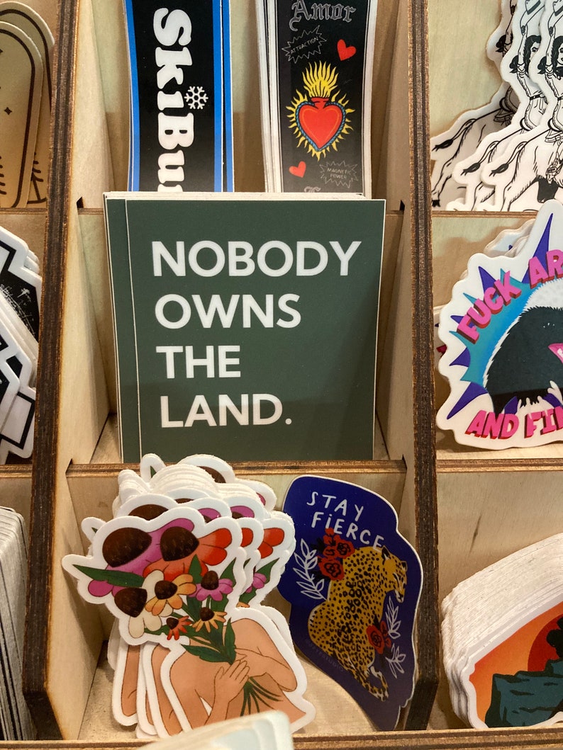 May include: A collection of stickers with various designs, including a green sticker with the text "Nobody owns the land." and a blue sticker with the text "Stay fierce" and a leopard illustration.