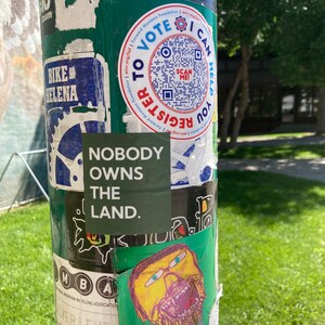 May include: A green pole with various stickers on it. One sticker says "Nobody owns the land." Another sticker has a QR code and says "To Vote Register".