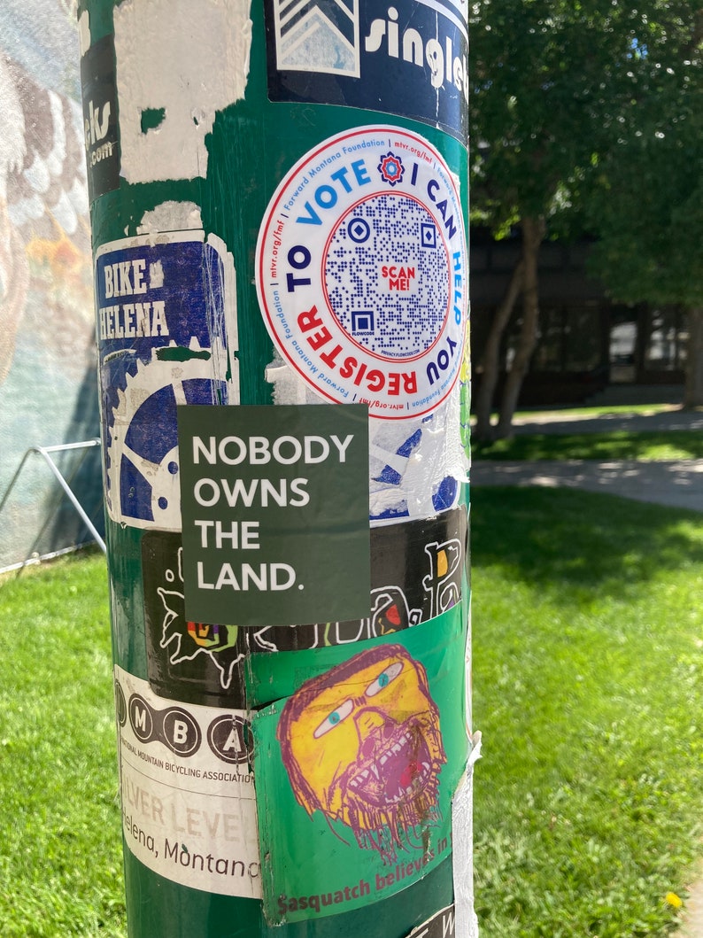 May include: A green pole with various stickers on it. One sticker says "Nobody owns the land." Another sticker has a QR code and says "To Vote Register".