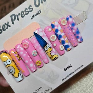 Puede incluir: Un conjunto de uñas postizas de lujo con diseños de personajes de dibujos animados. Las uñas son principalmente rosas, con lunares blancos, e incluyen imágenes de Homer y Marge Simpson. Algunas uñas tienen adornos de rosquillas y gemas. El texto "Sex Press On Nails" es visible.