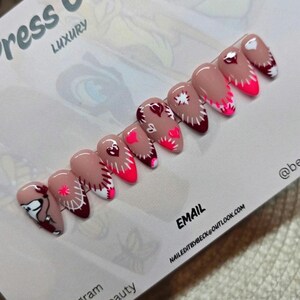 Puede incluir: Un conjunto de uñas postizas en forma de almendra con base nude y varios diseños. Los diseños incluyen corazones rojos, costuras blancas y puntas rosa neón. La tarjeta dice "Press On Nails LUXURY" e incluye información de Instagram y correo electrónico.
